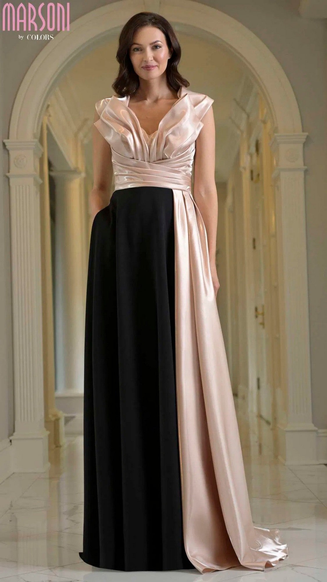 A woman stands in an elegant hallway wearing the Marsoni M345 long formal dress in Black Champagne, featuring a v-neckline and a stylish black skirt.